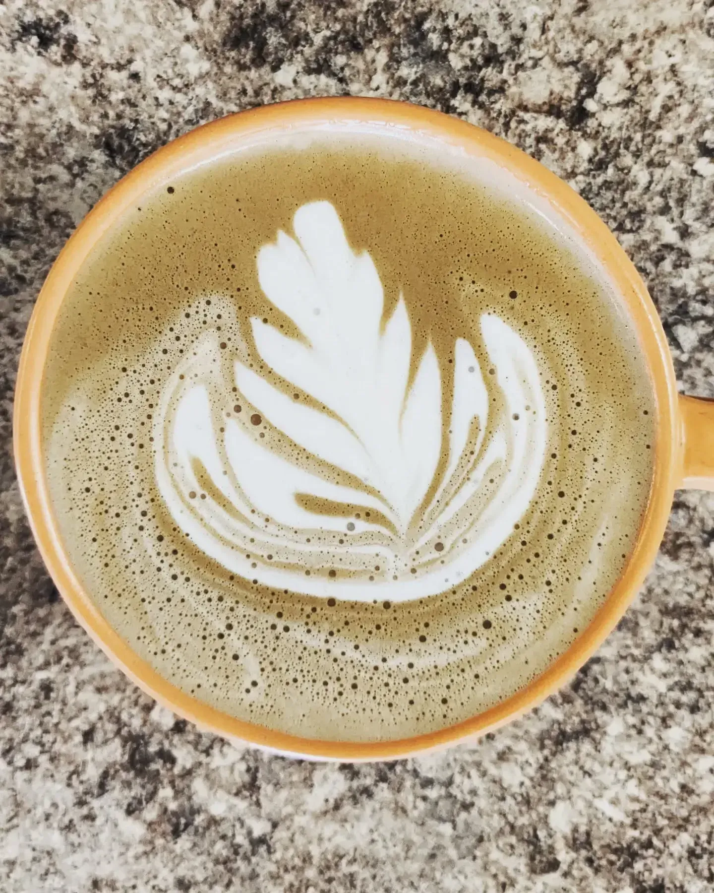 Latte in an orange mug with white leaf art on textured surface.