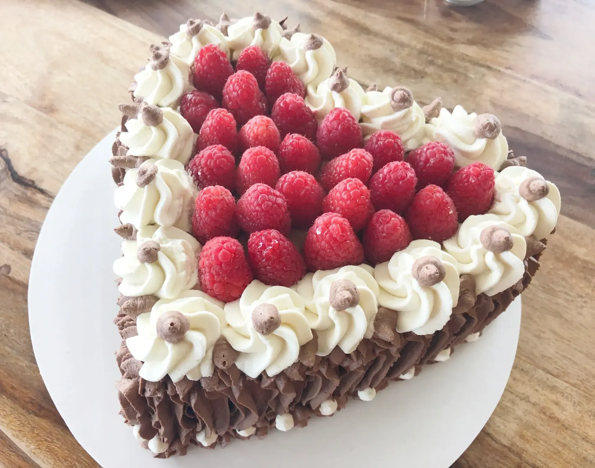 Heart-shaped chocolate cake decorated with white frosting swirls, fresh raspberries, and chocolate chips.