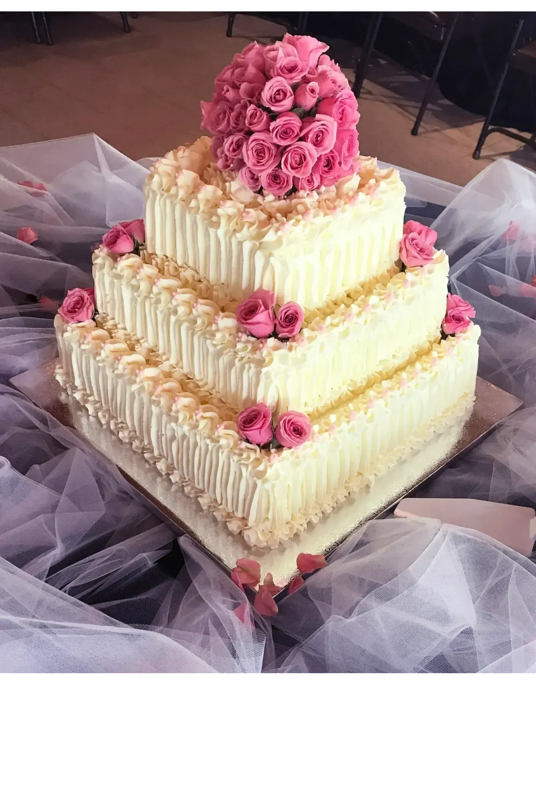 Three-tiered square cake decorated with white frosting and pink roses on a reflective surface.