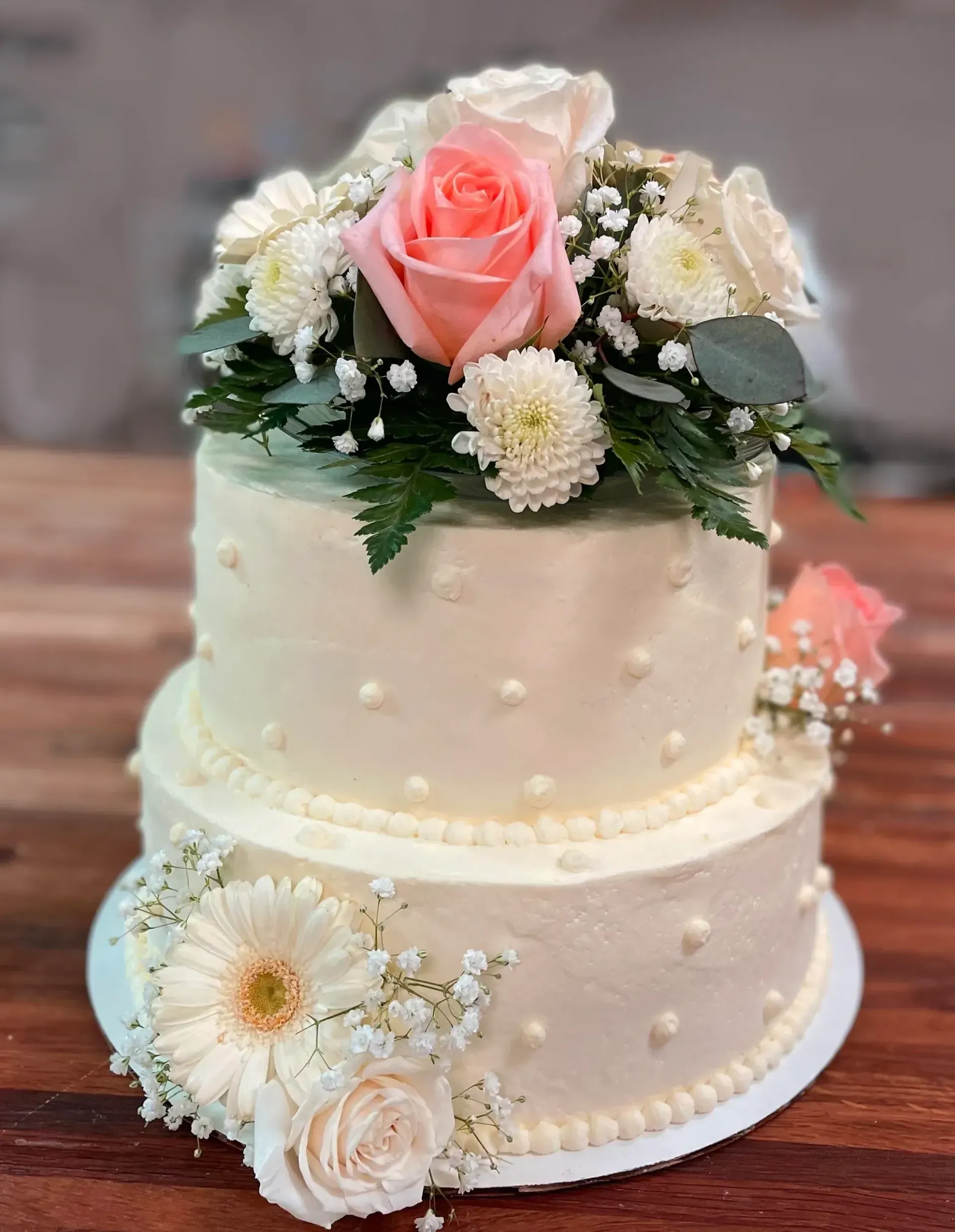 Two-tiered white cake, decorated with pink and white roses, chrysanthemums, and baby's breath.