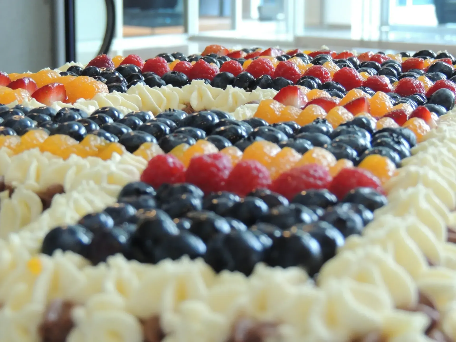 Close-up of a rectangular cake, decorated with whipped cream, blueberries, raspberries, mandarin oranges, and strawberries.