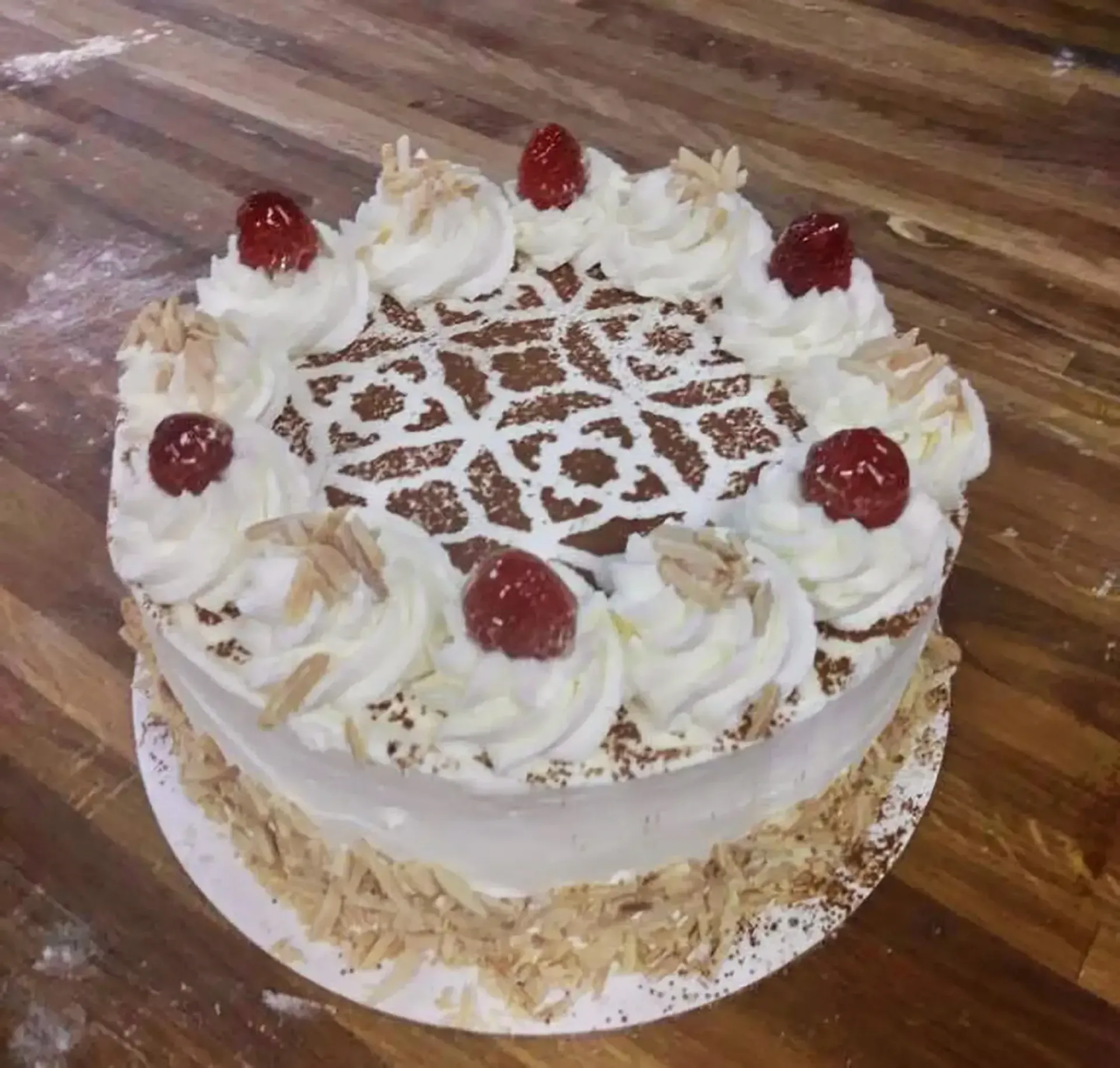 Cake with white frosting, red cherries, almond slices, and decorative icing on a wooden table.