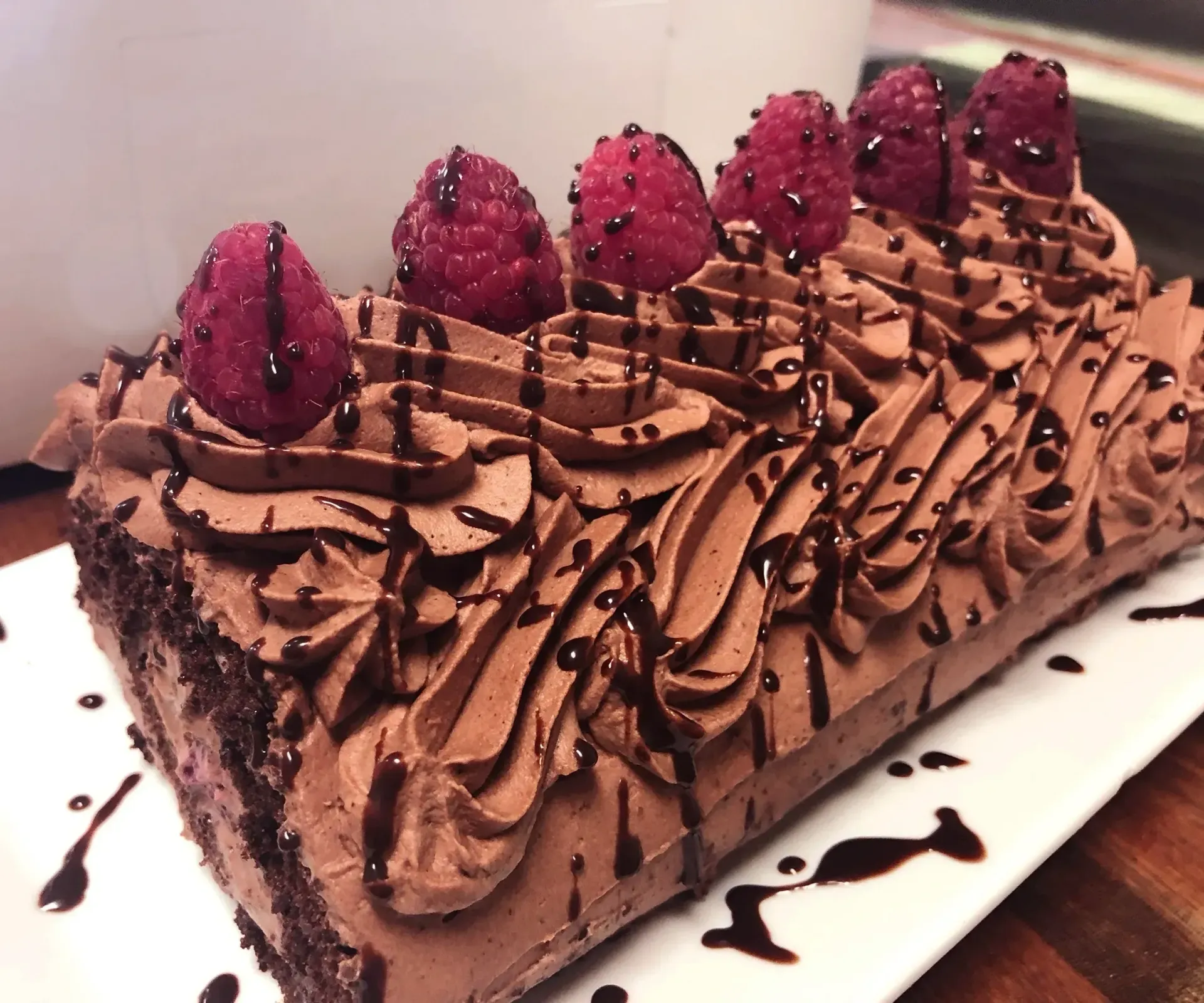 Chocolate cake log decorated with chocolate frosting, raspberry and chocolate drizzle on a white plate.