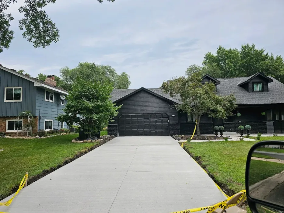 New concrete driveway leading to a dark house with trees and adjacent blue house. Construction tape borders the driveway.