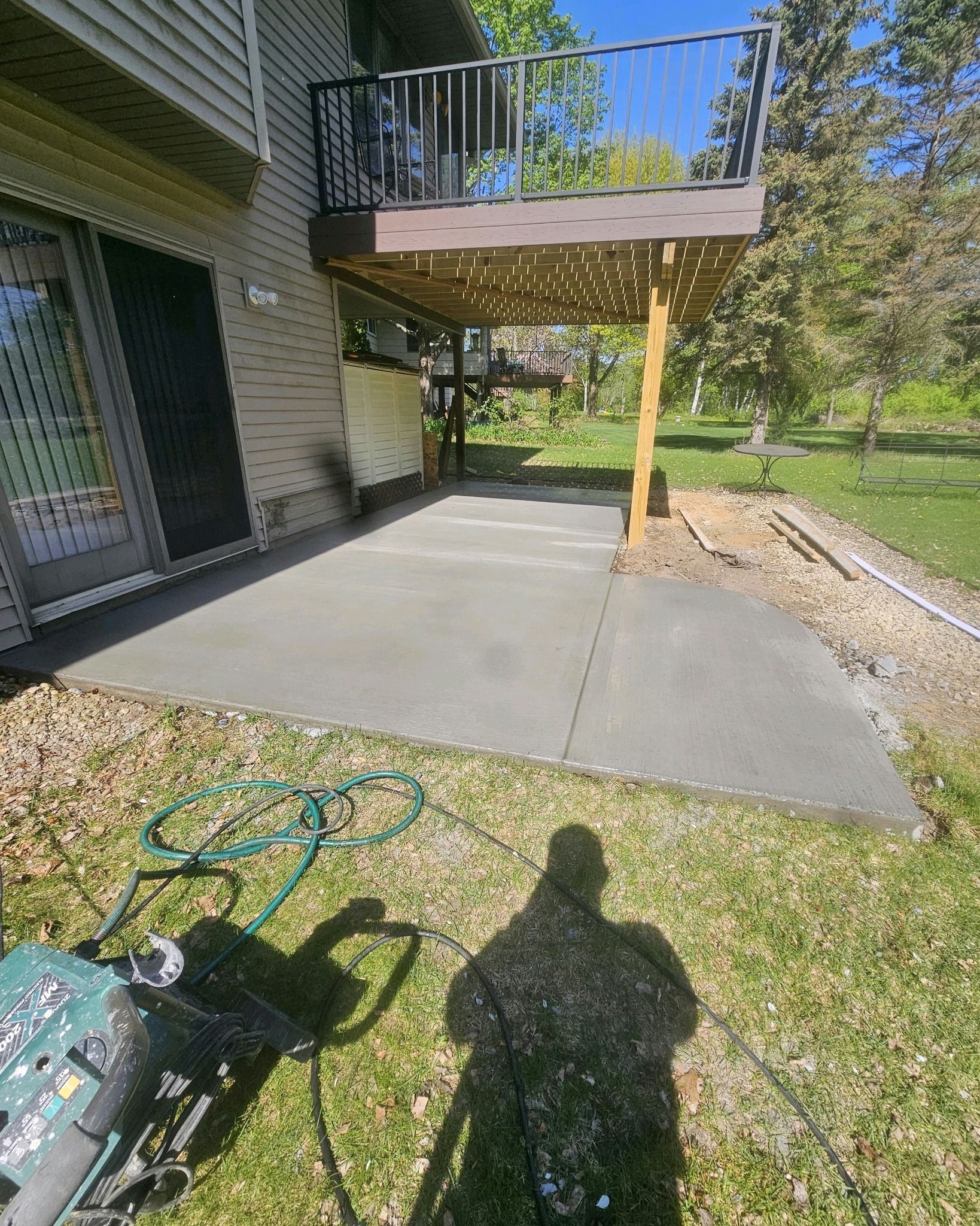 A concrete patio with a deck overhead, grass, and a person's shadow.