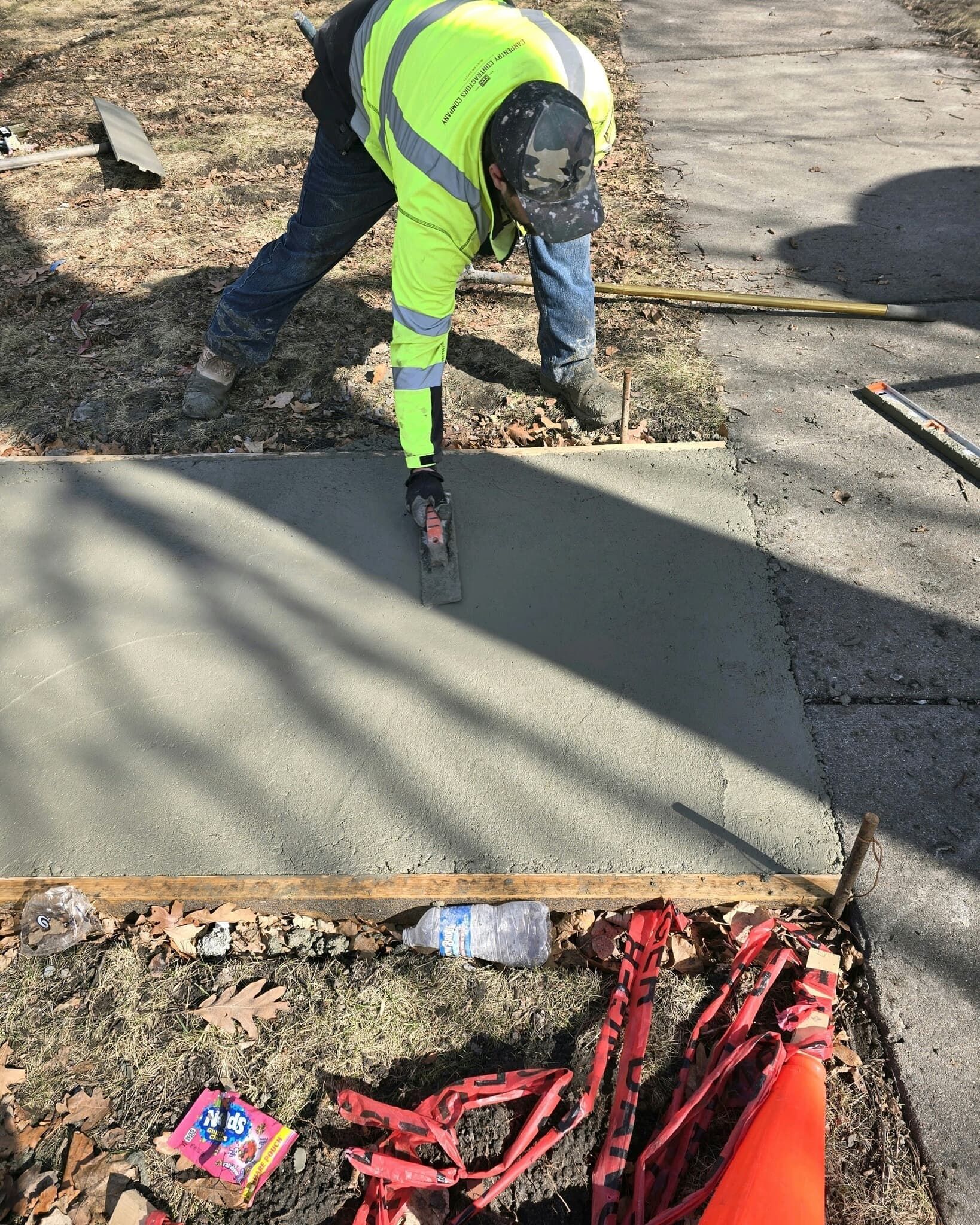 Construction worker in reflective vest smoothing fresh concrete sidewalk.