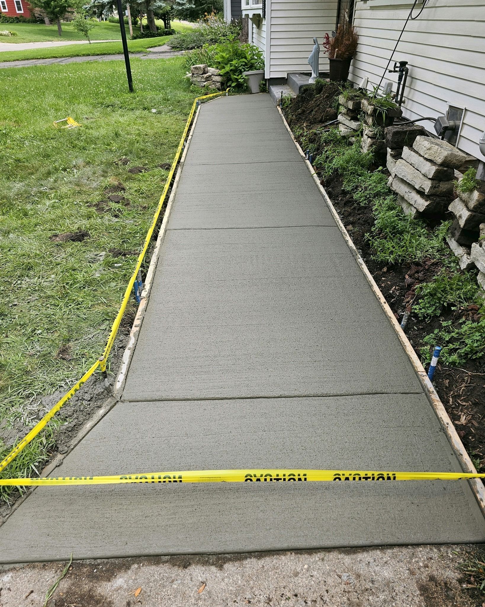 Freshly poured concrete sidewalk with yellow caution tape, next to grass and a building.