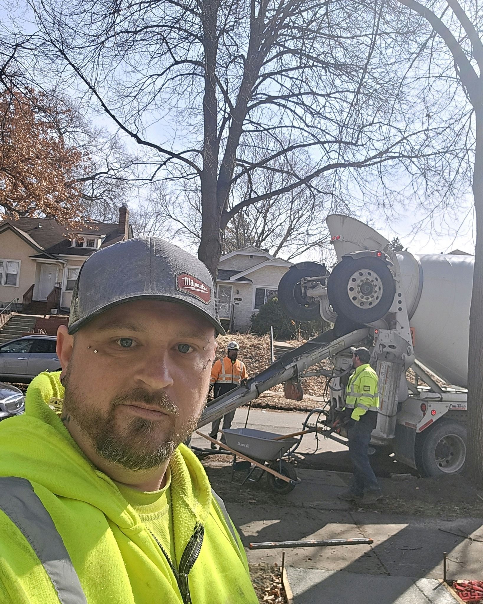 Man in neon yellow jacket, baseball cap, with cement truck pouring concrete.