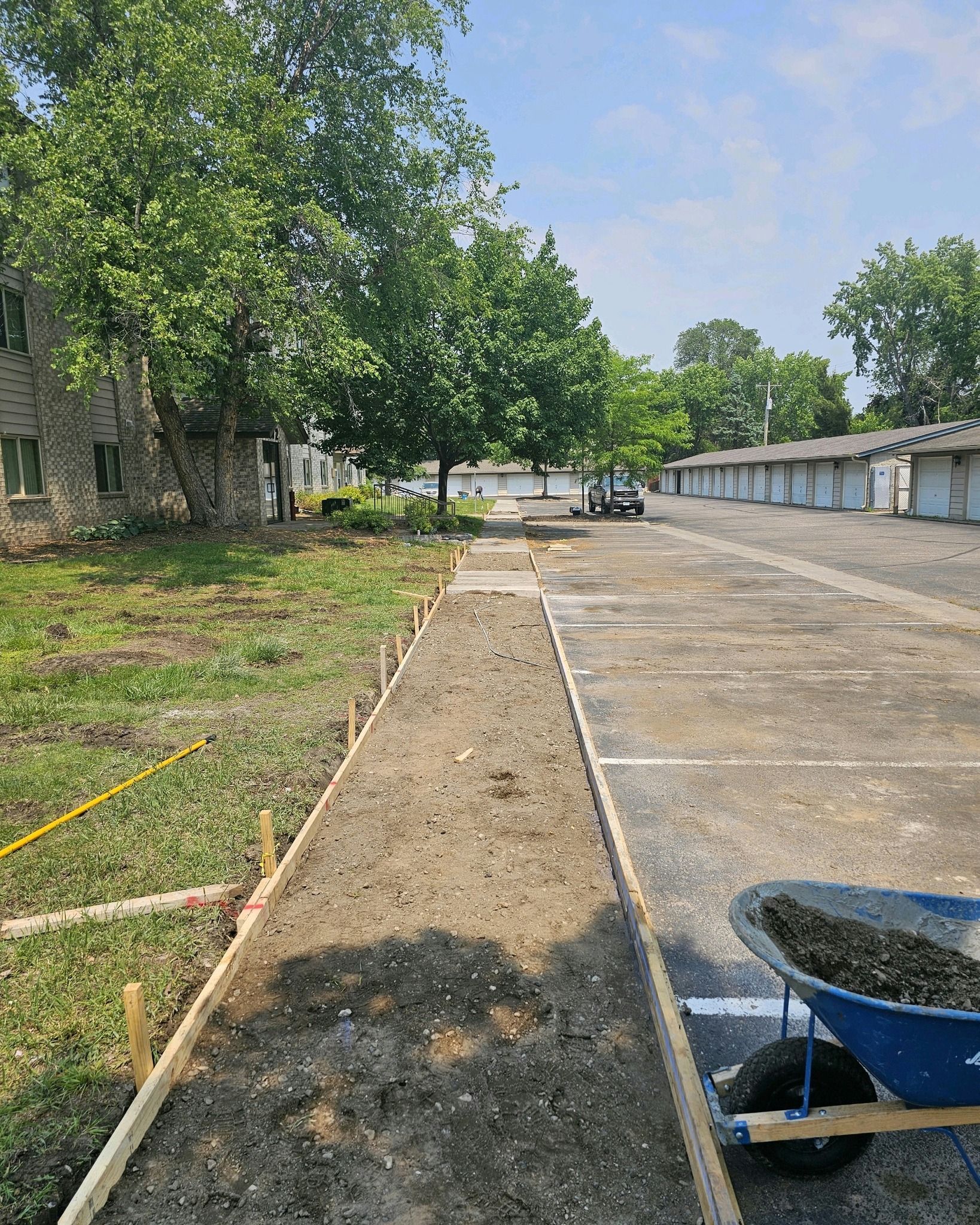 Concrete path under construction next to a parking lot and grass. Blue wheelbarrow of gravel.