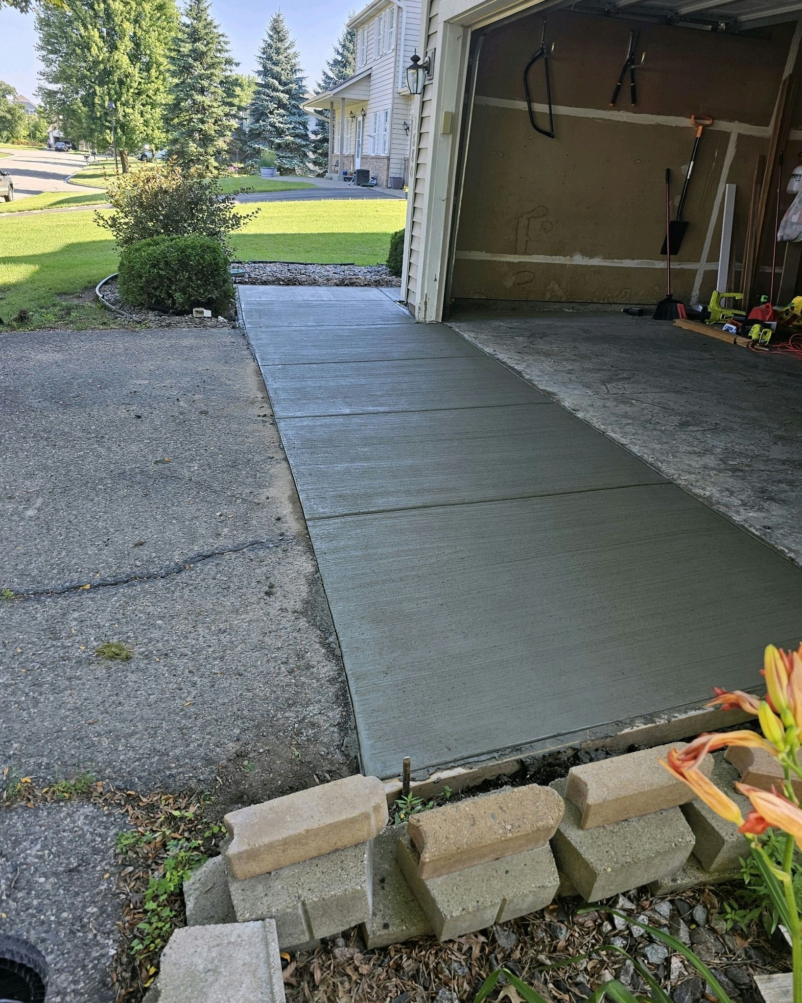 Newly poured concrete walkway leading to a garage. Adjacent to existing driveway, a small garden with stone bricks is visible.