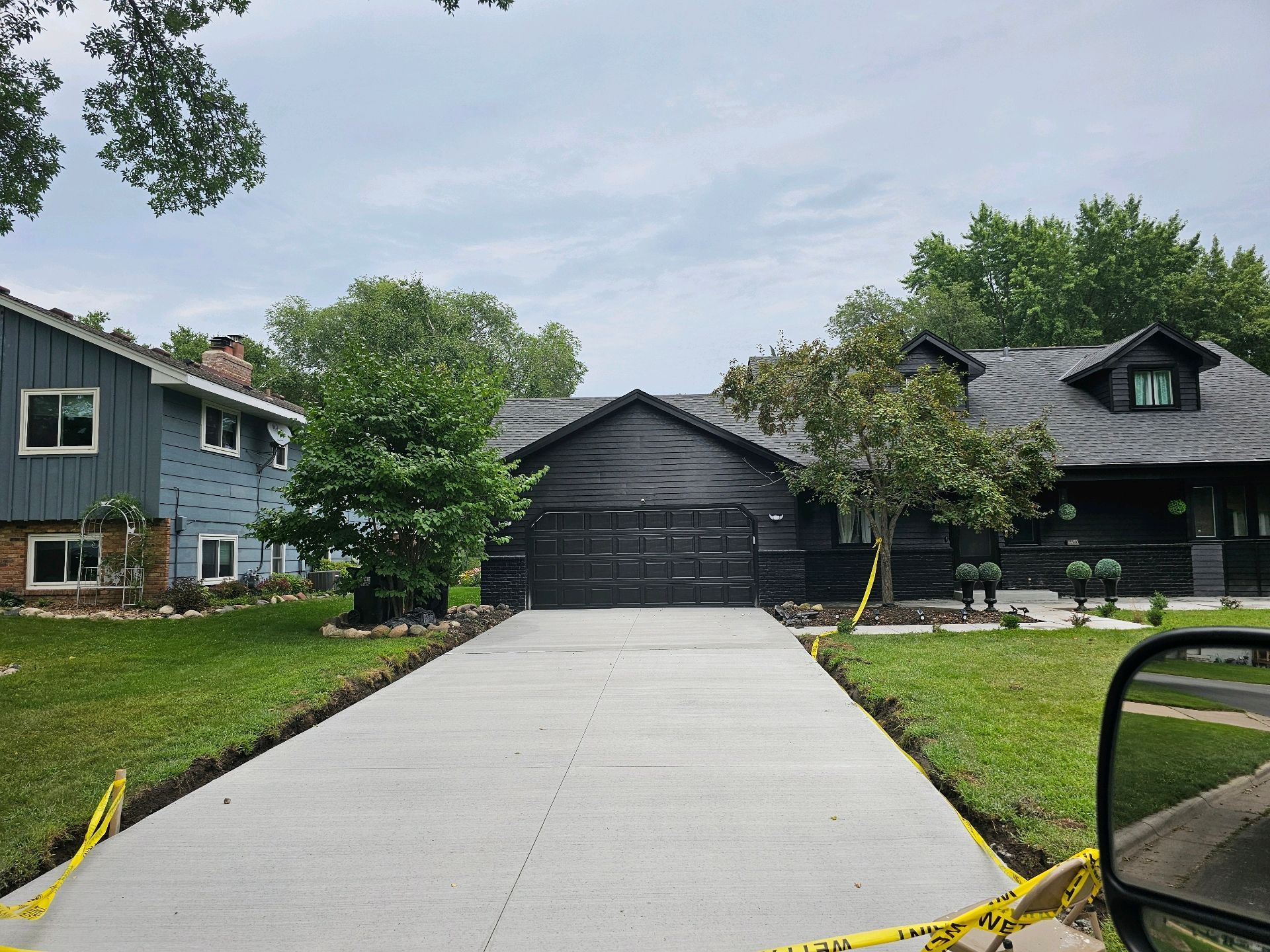 A newly poured concrete driveway leads to a dark-colored house with trimmed landscaping; yellow caution tape is present.