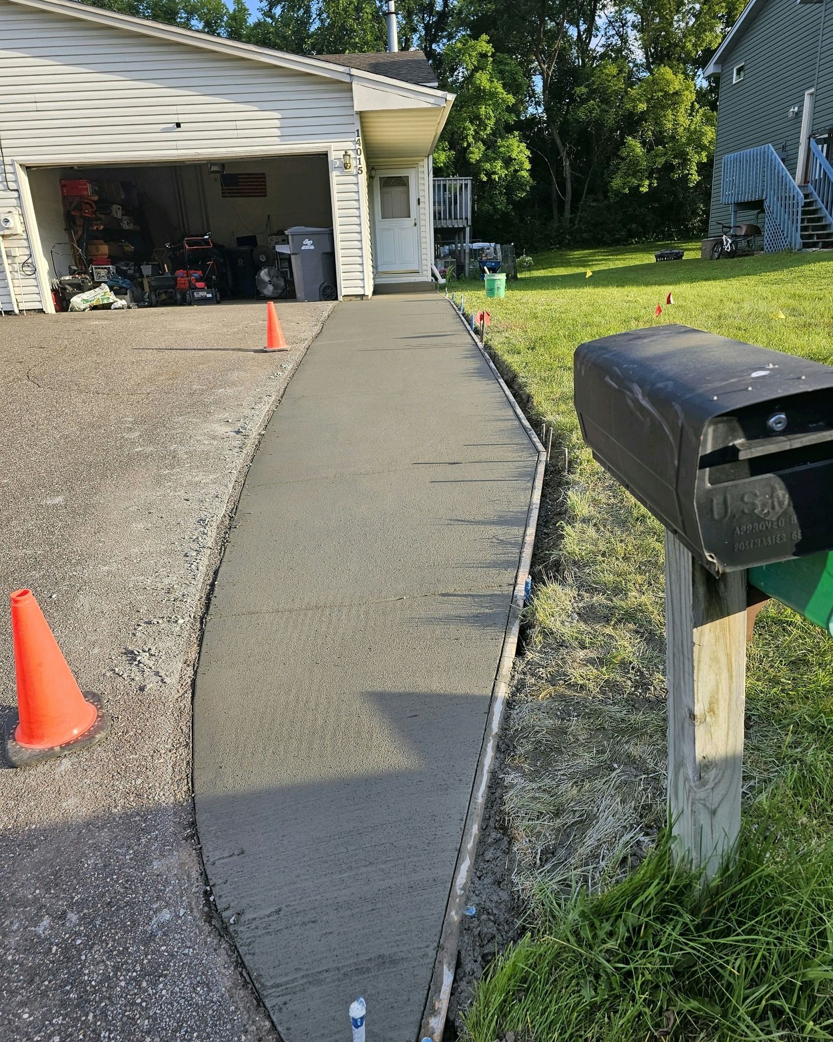 New concrete walkway next to a driveway and grassy yard, with an orange cone and mailbox.