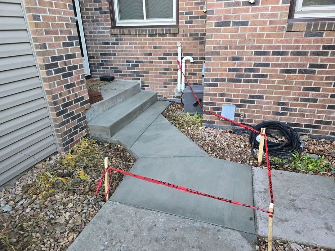 Newly poured concrete walkway and steps, red tape barrier, brick building exterior.