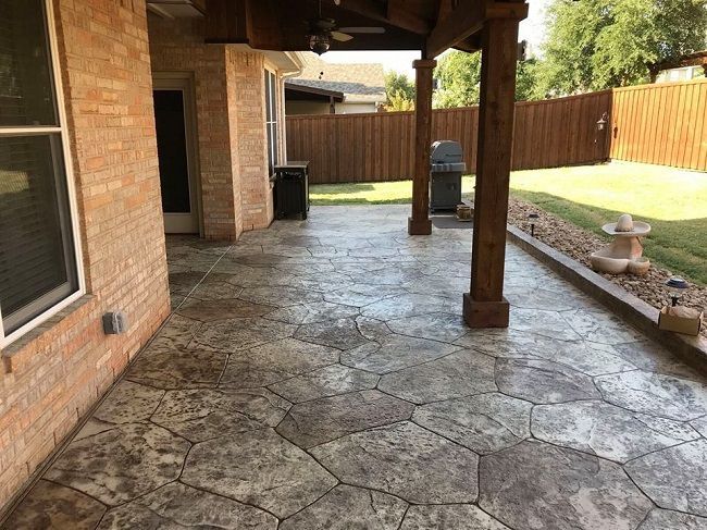 Patio with textured stone-like concrete flooring and a wooden pergola, next to a brick building.