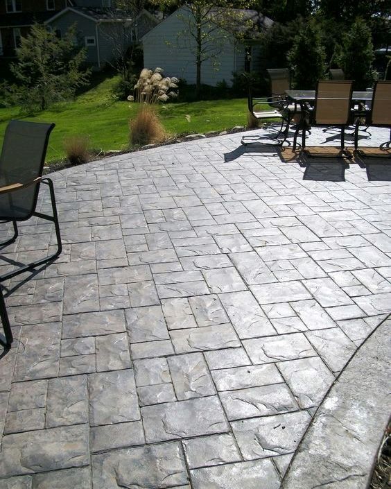 Stamped concrete patio, gray with stone pattern, chairs and table, green lawn and shed in the background.