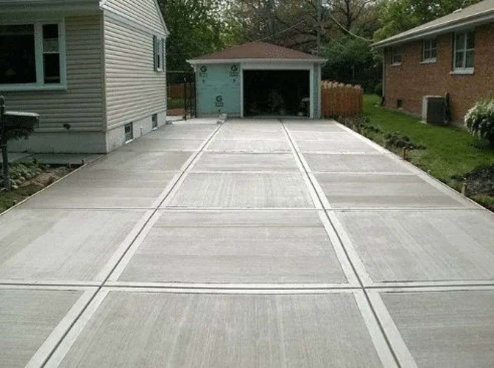 Concrete driveway with rectangular sections leading to a garage. Houses are on either side with green grass.