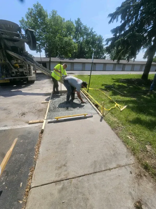 Workers pouring concrete for a sidewalk next to a parking lot under a sunny sky.