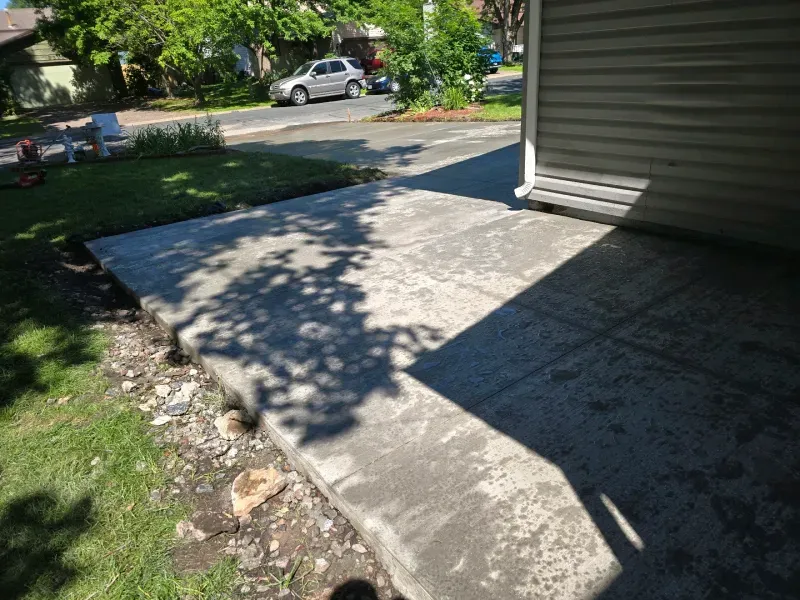 Concrete patio with tree shadow, adjacent to house with siding, next to grass and street.