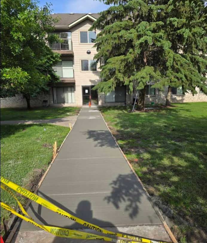 Newly poured concrete sidewalk leading to an apartment building entrance, yellow caution tape on the edges.