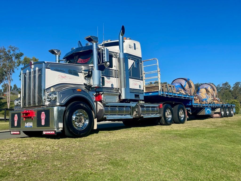 Shiny white and chrome semi-truck with flatbed trailer hauling large cable spools on a grassy field — ECC Heavy Haulage in Ormeau, QLD