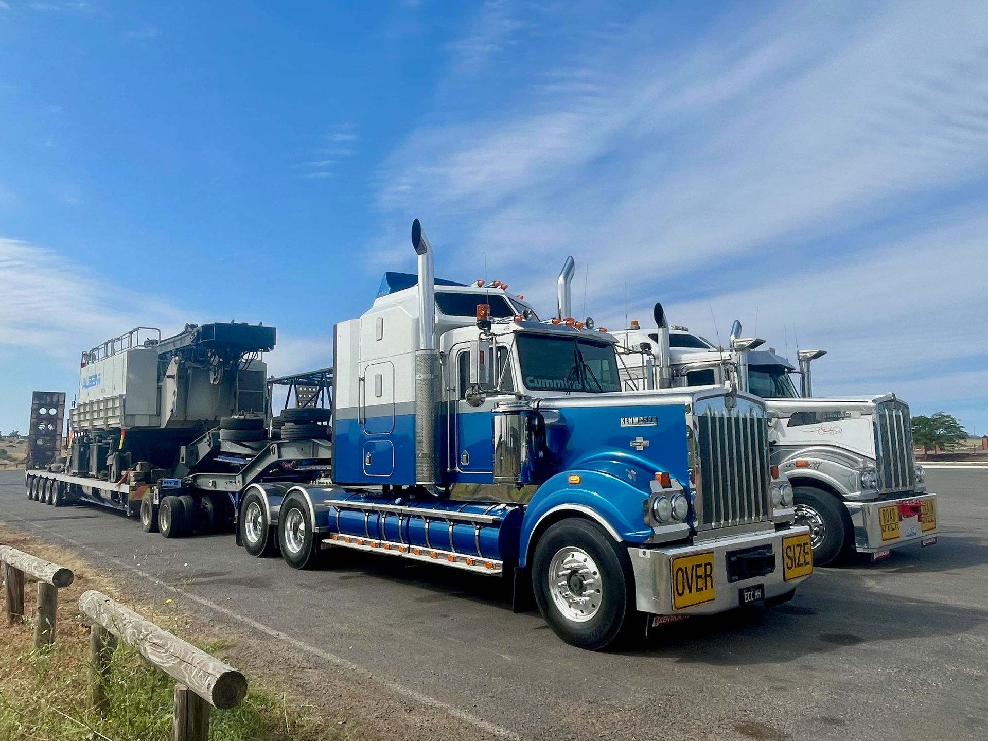Two semi-trucks, one blue and white, hauling heavy equipment on a sunny day — ECC Heavy Haulage in Ormeau, QLD