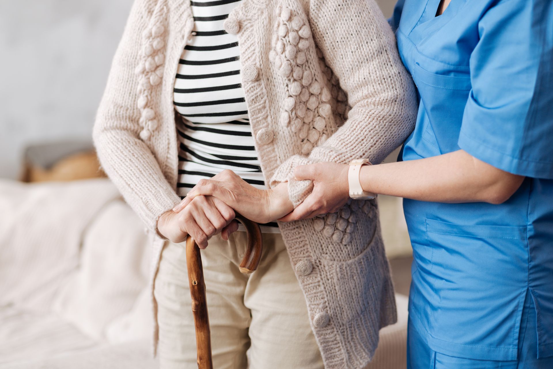 A caregiver in blue scrubs supports a person holding a wooden cane while they stand.