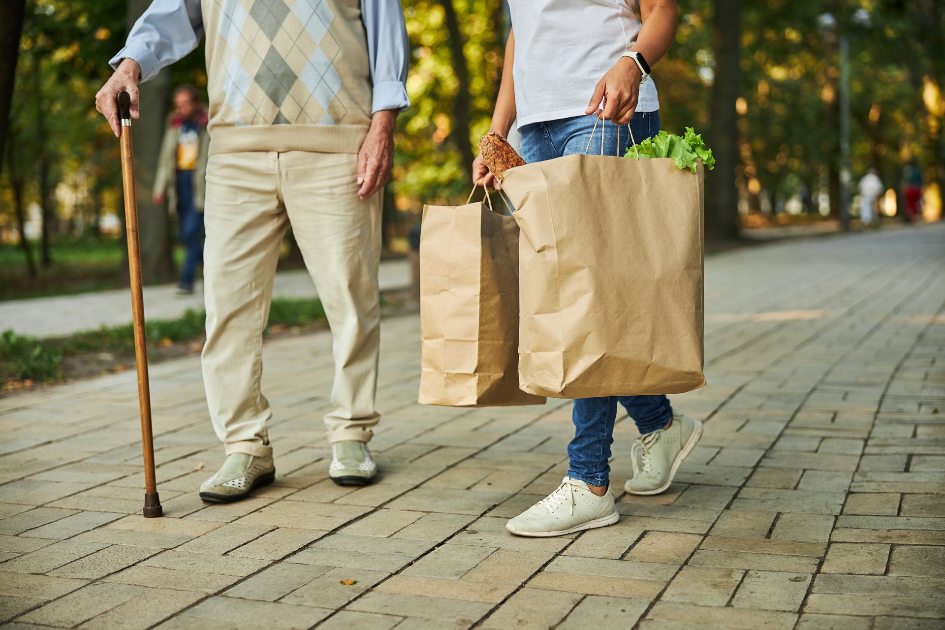 Two people walk together on a paved park path, carrying brown paper grocery bags. One person uses a wooden cane. Two people walk together on a paved park path, carrying brown paper grocery bags. One person uses a wooden cane.
