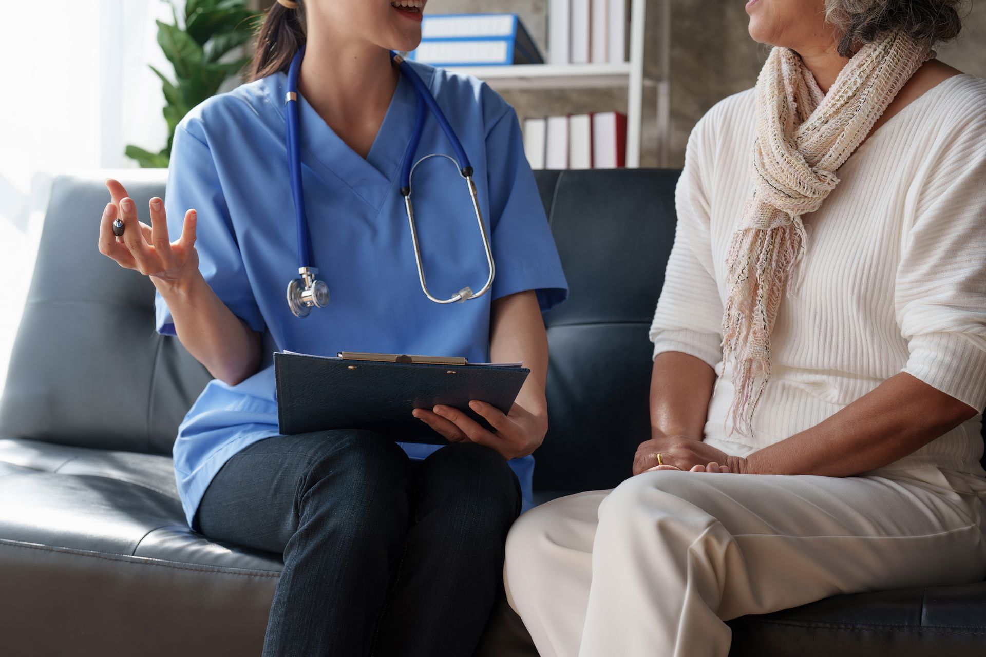 A medical professional in blue scrubs sits on a couch, holding a clipboard and talking with a patient wearing a scarf.