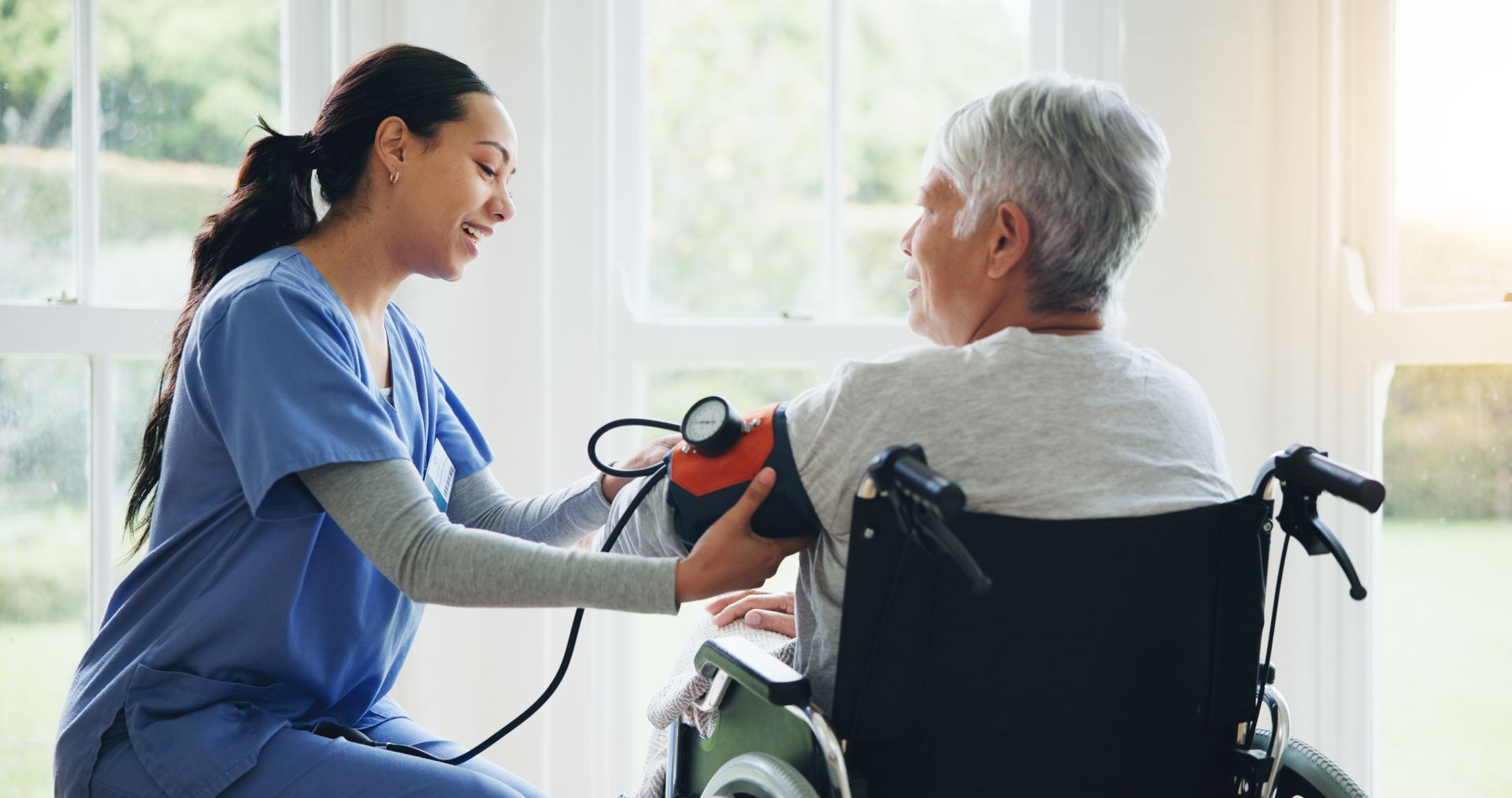 A caregiver in blue scrubs takes the blood pressure of a person seated in a wheelchair by a bright window. A caregiver in blue scrubs takes the blood pressure of a person seated in a wheelchair by a bright window.
