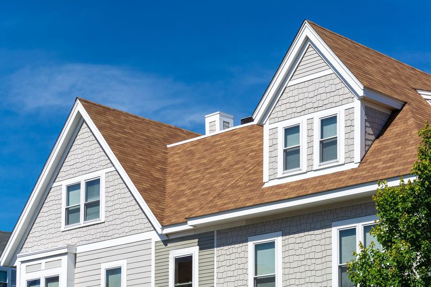Tan shingled roof with gray siding, dormer windows, and a blue sky.