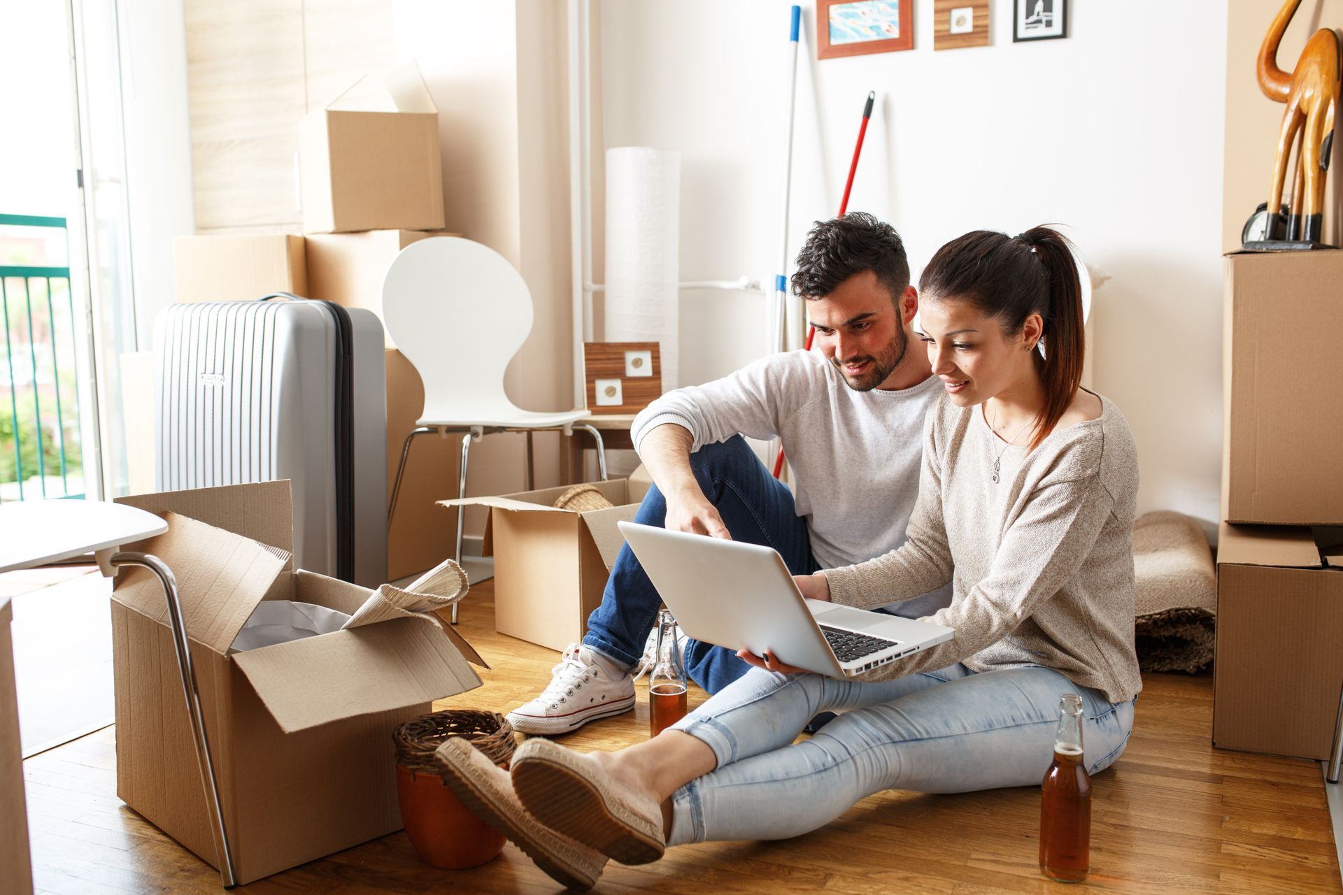 Couple sits on floor with laptop, surrounded by moving boxes in a bright room.