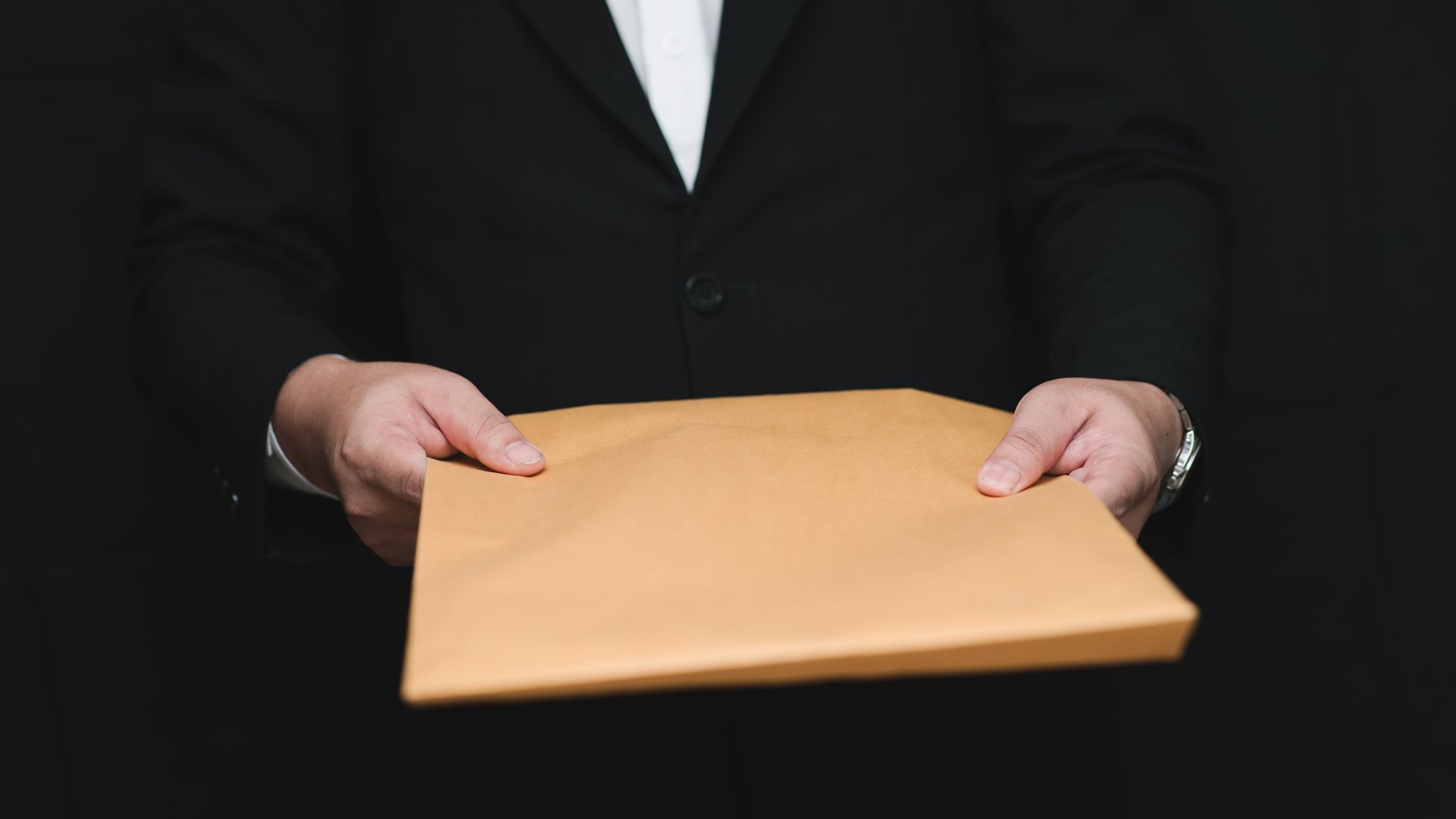 Person in suit holding out a brown envelope against a black background.