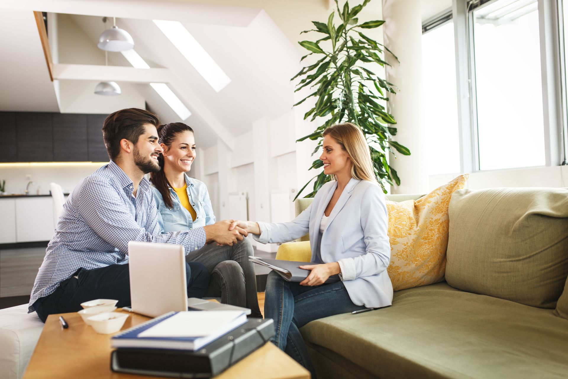Couple shaking hands with a realtor on a couch; laptop and documents on the table.