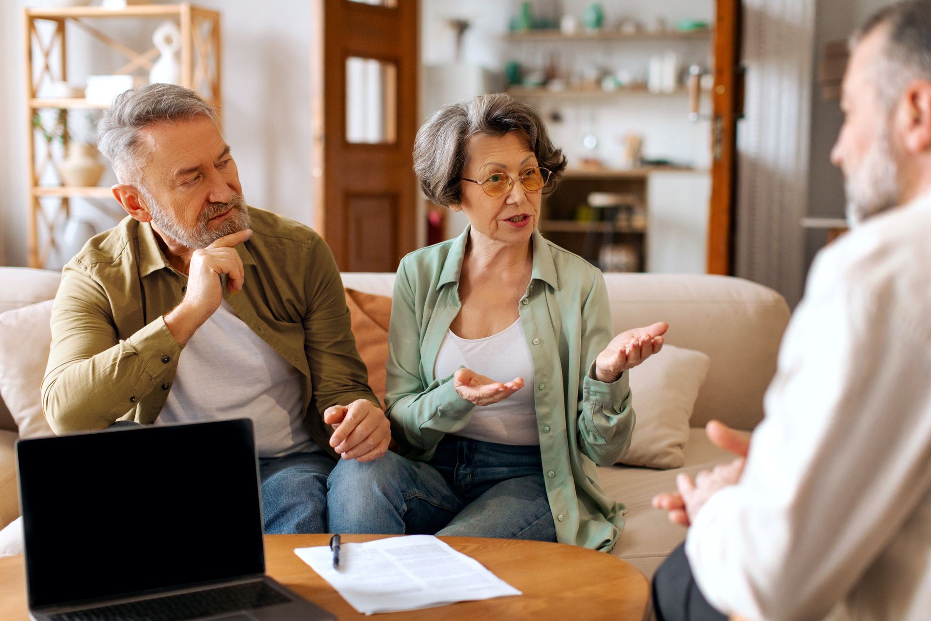 Couple consults with a professional in a living room, discussing paperwork, laptop on table.