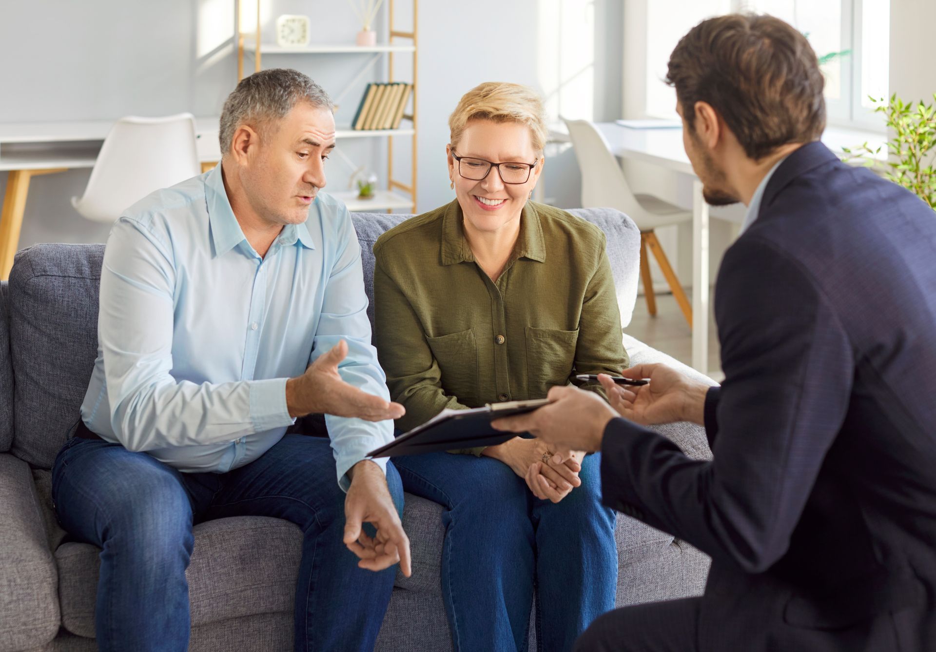 Couple on a couch consults with a person holding a clipboard; indoor setting.