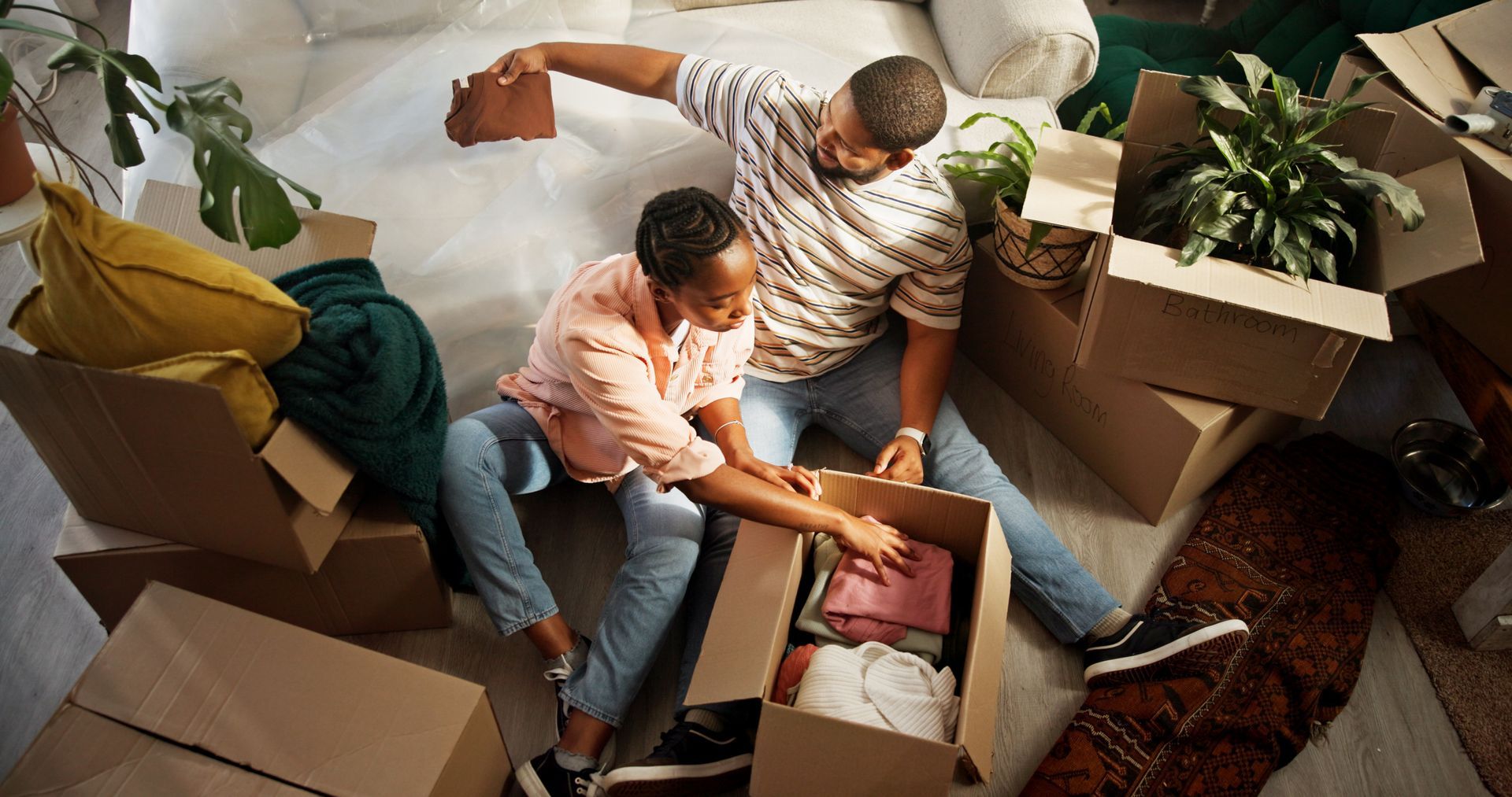 Two people unpacking boxes, surrounded by moving supplies, in a room with plants.