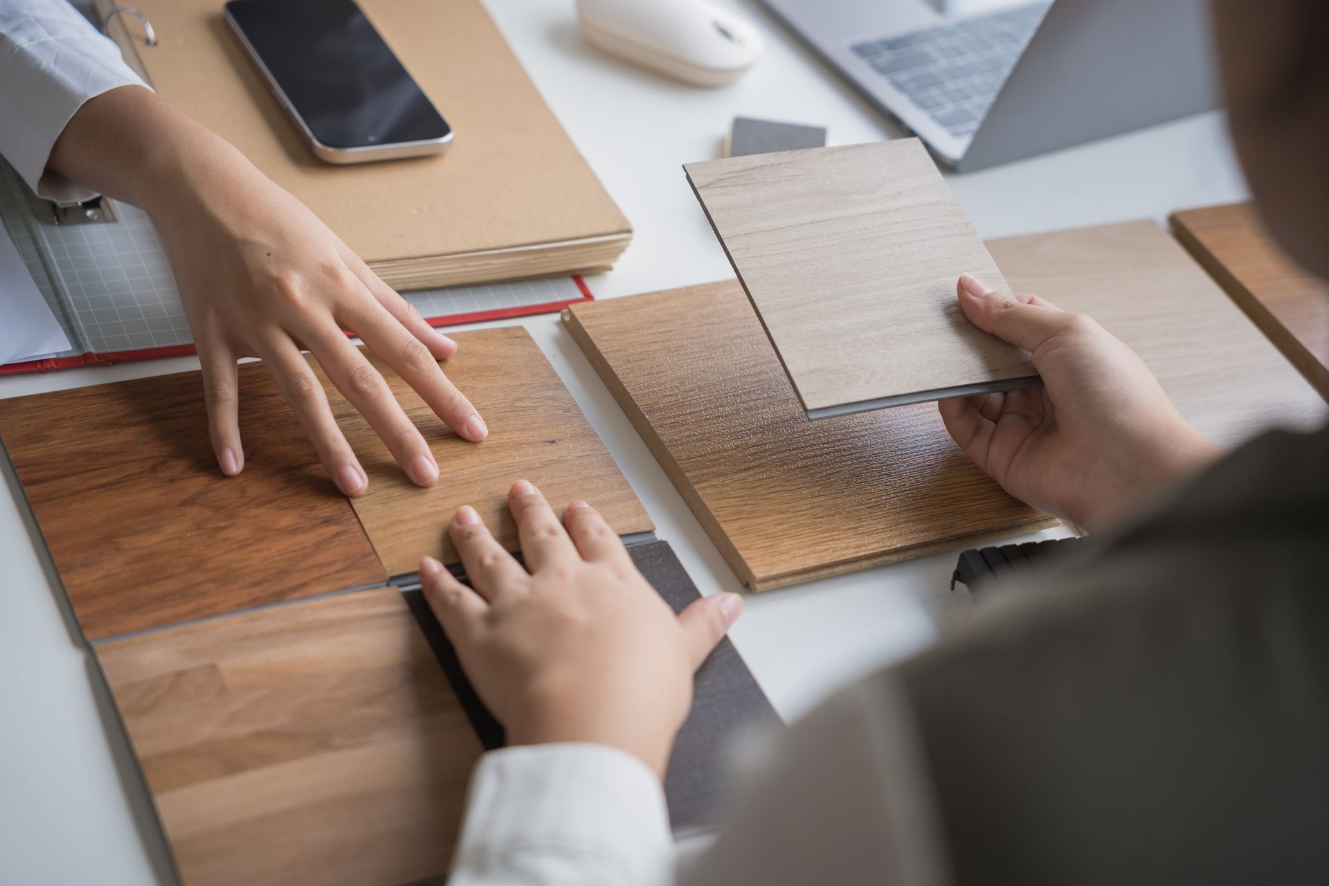 Hands comparing wood flooring samples on a white table. Laptop and phone nearby.