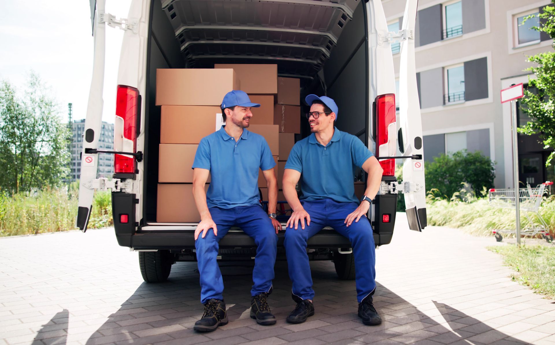 Two movers in blue uniforms, resting in back of van packed with boxes.