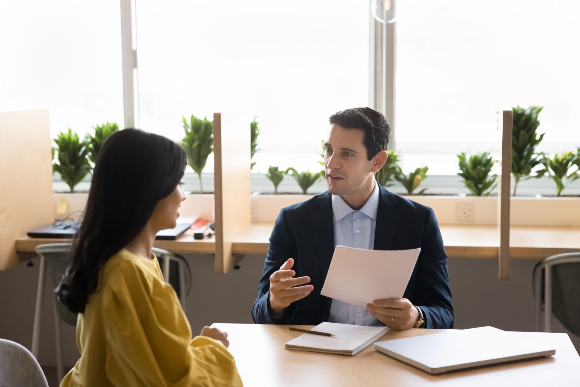 Man in suit reviews documents with woman in yellow top, sitting at a table with a window background.