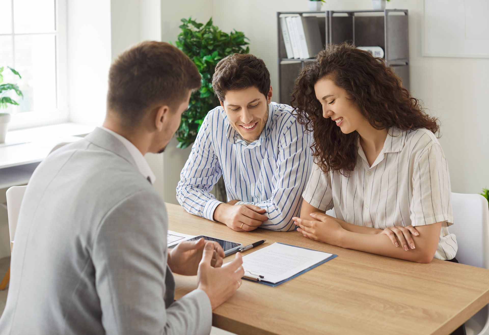 A financial advisor, a man in a gray suit, consults with a smiling couple at a desk with paperwork in a light-filled office.