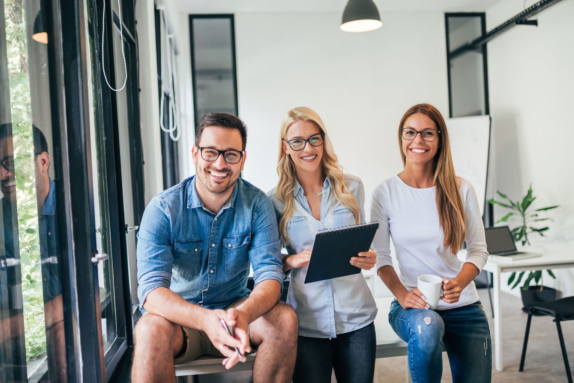 Three people, one with a clipboard, smiling in an office setting with a window.