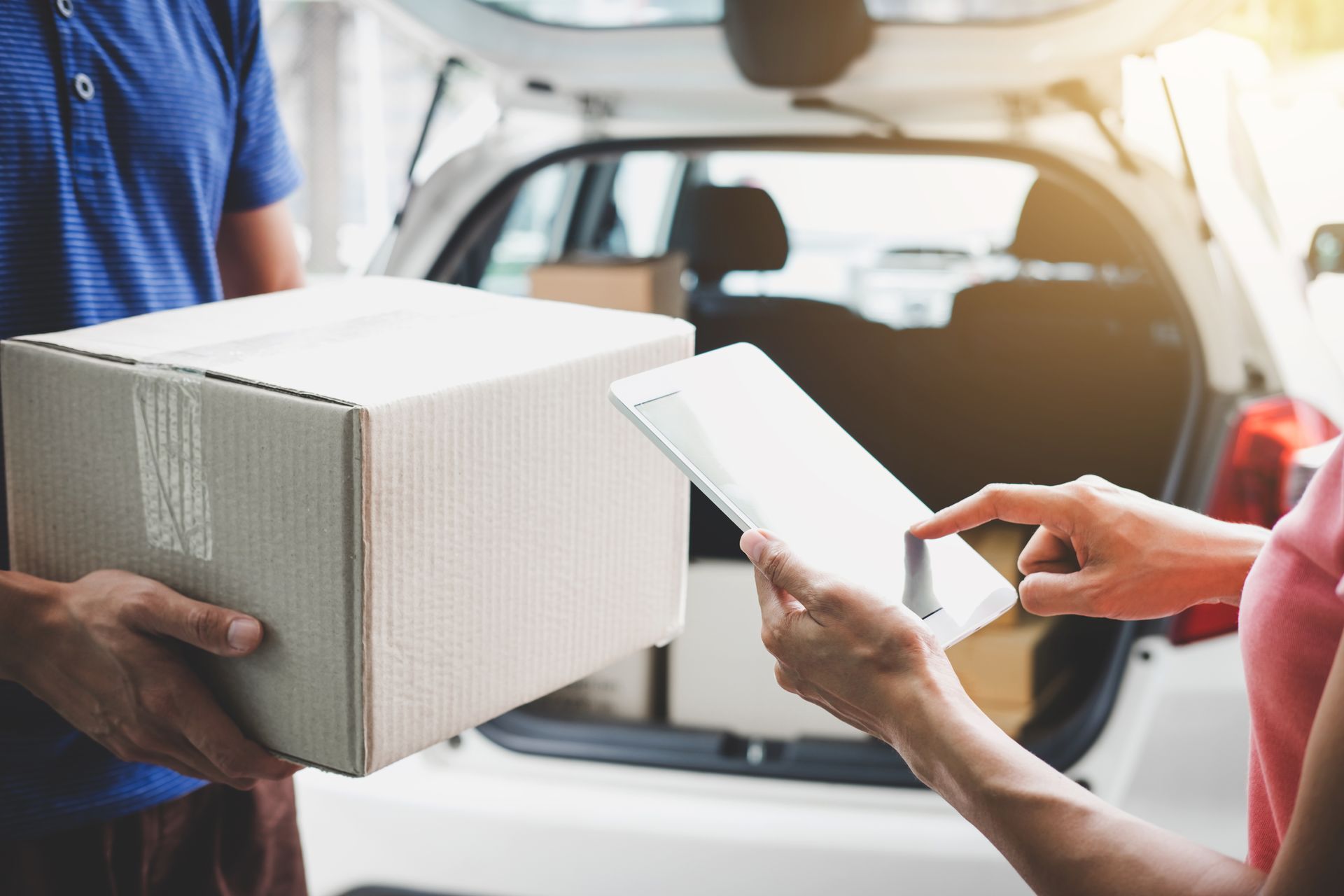 Delivery person handing a cardboard box to a person signing a digital tablet by a car trunk.
