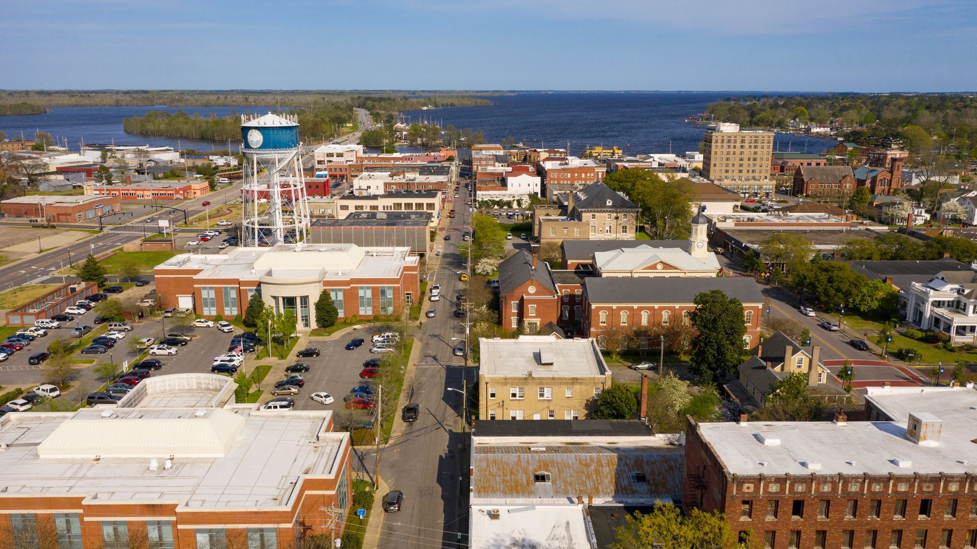 Aerial view of a downtown area with buildings, roads, and a large body of water in the background under a blue sky.