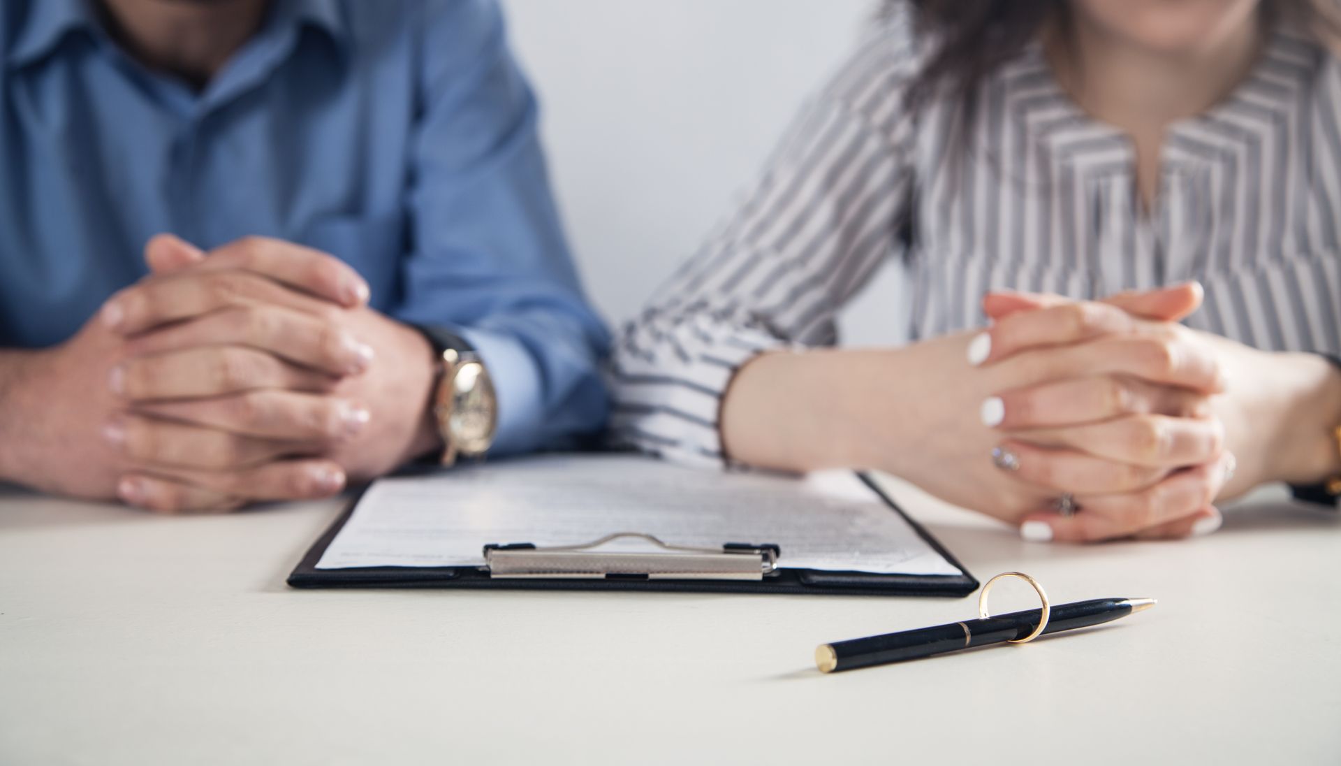 Two people with clasped hands sit at a table with a clipboard and pen.