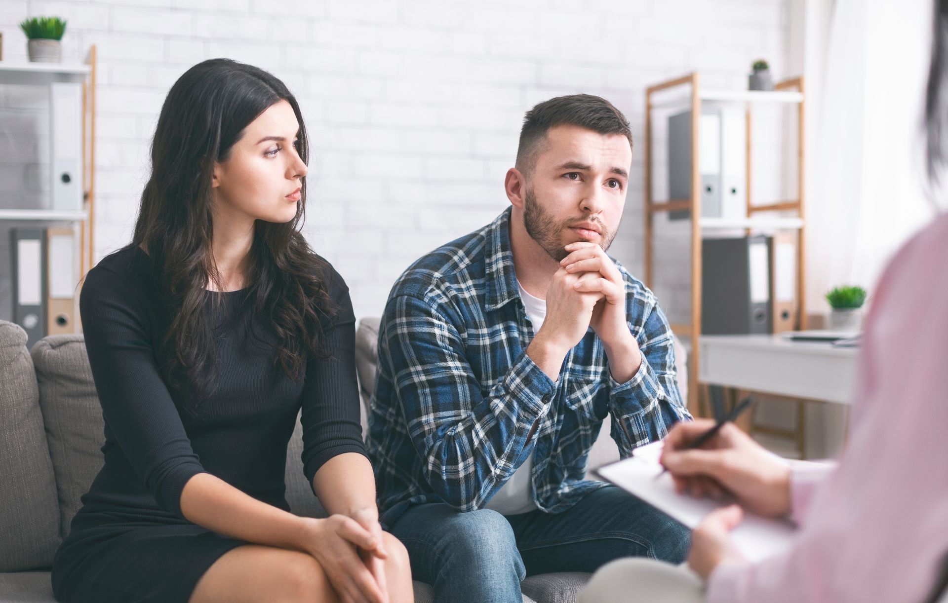 Couple sitting on a couch, looking somber during a therapy session. A therapist takes notes.