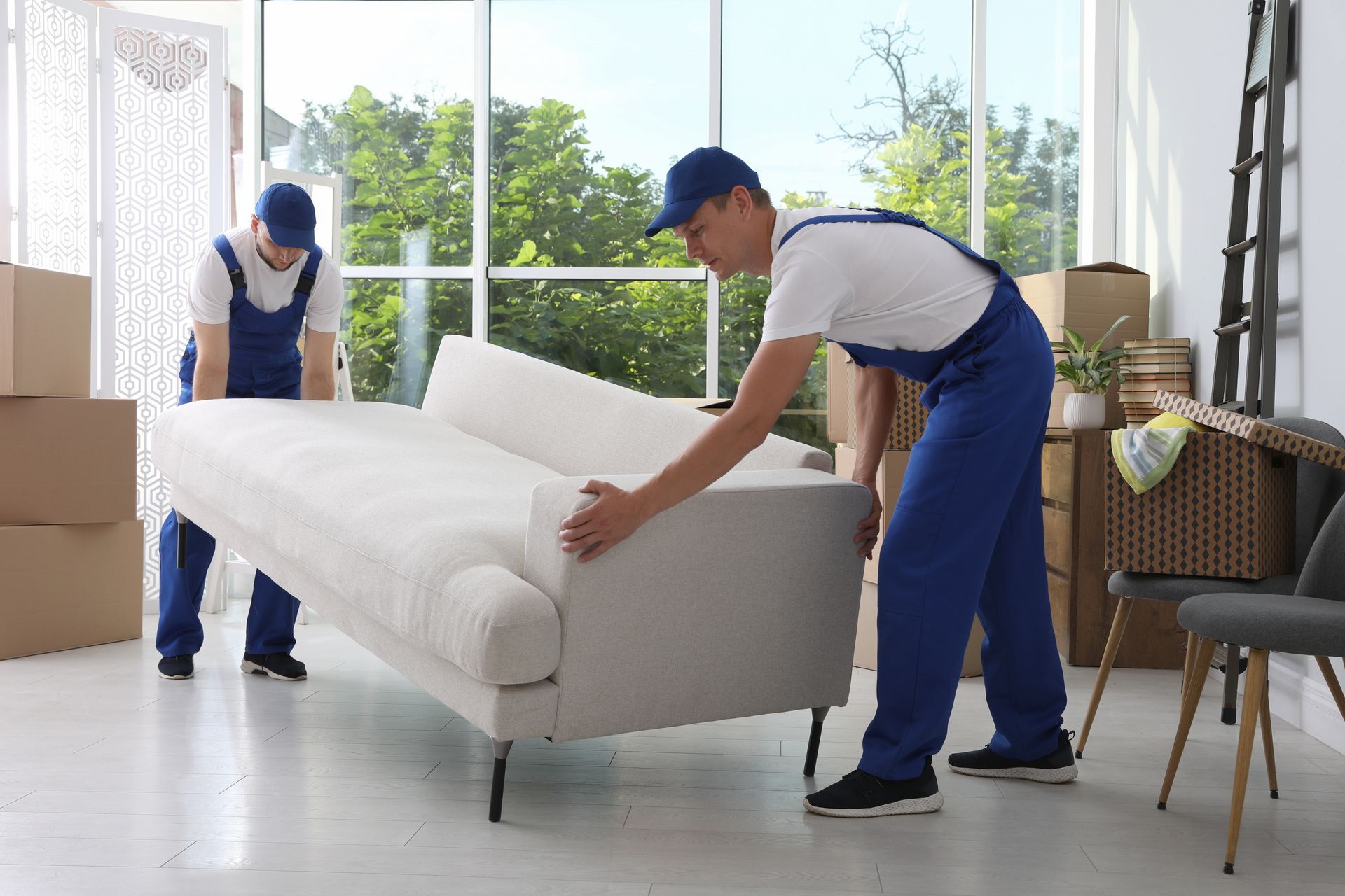 Movers in blue overalls carry a white couch in a bright room with boxes.