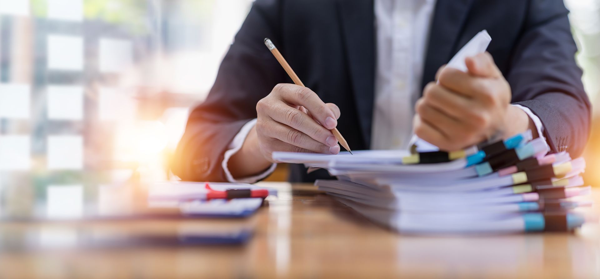 Person in a suit writing on paper with a pencil at a desk with stacked files.