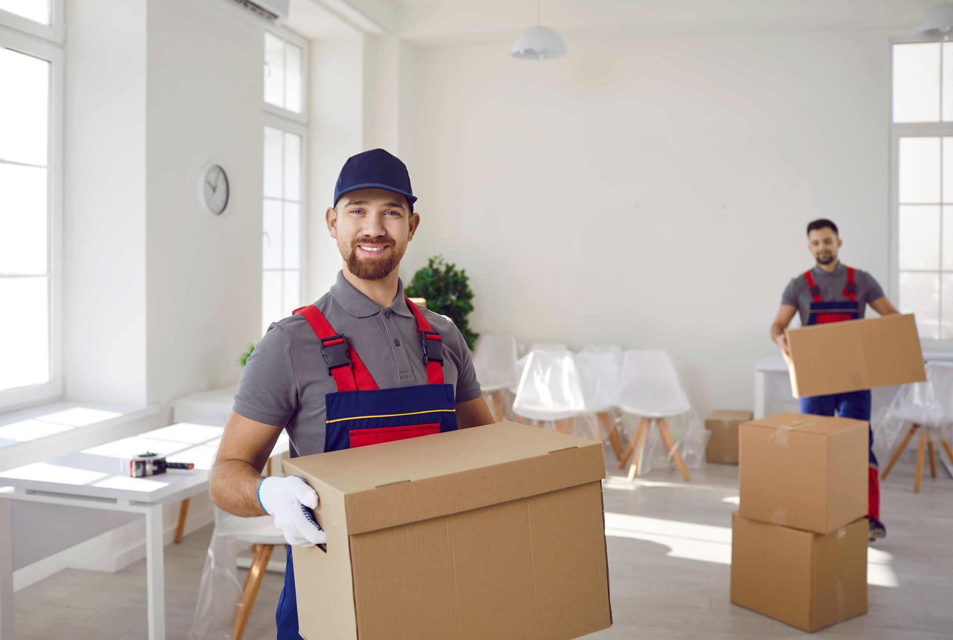 Two movers in a bright room holding cardboard boxes, one smiling at the camera.