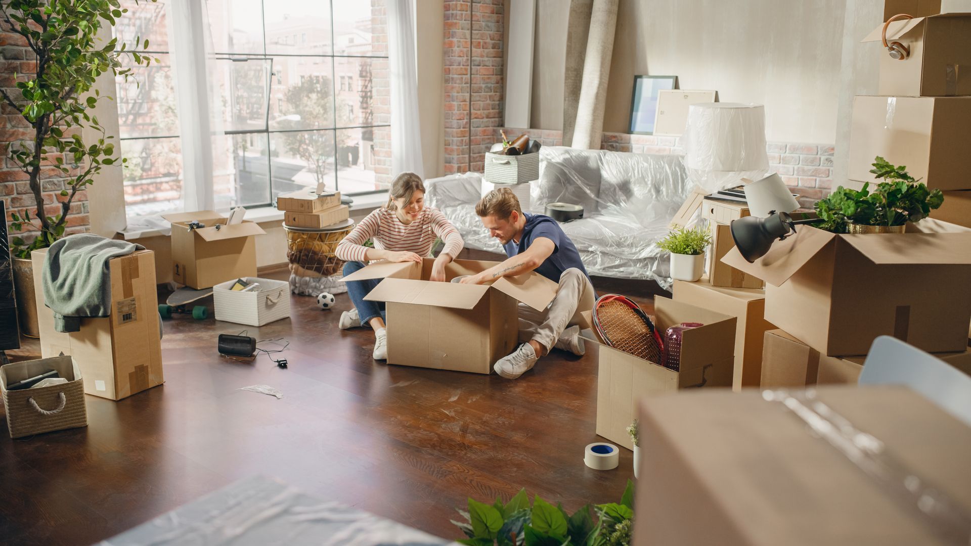 Two people unpacking boxes in a room with packed furniture, moving day.