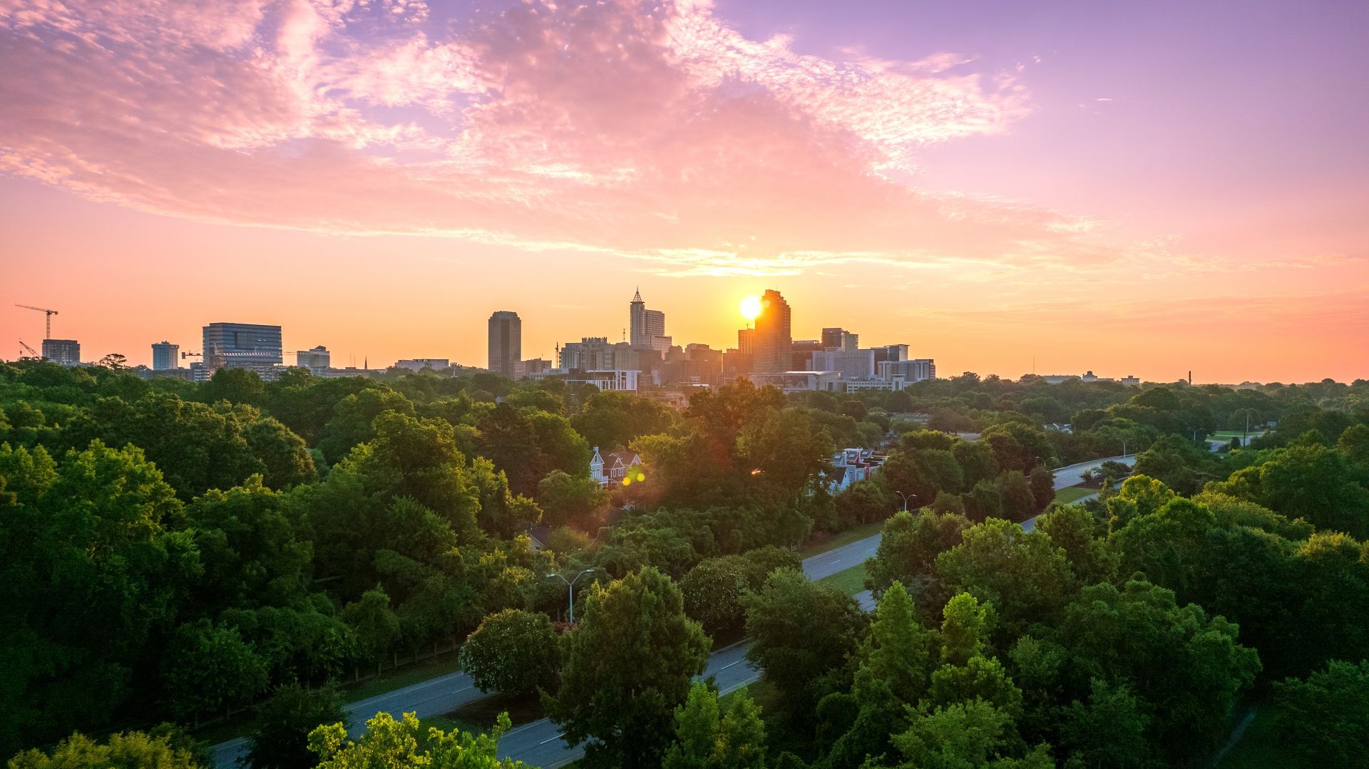 Sunrise over Raleigh, North Carolina skyline, viewed above lush green treetops, with pink and orange sky.